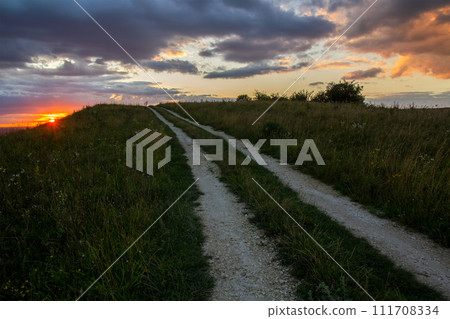 Dirt road among dark fields at sunset with dramatic cloudscape. 111708334