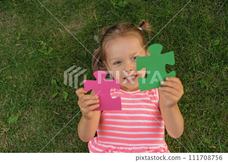 The child holds two large pieces in the shape of a puzzle lying on the grass. Intellectual development and acquisition of non-standard thinking skills The child holds two large pieces in the shape of a puzzle lying on the grass. Intellectual development and acquisition of non-standard thinking skills 111708756