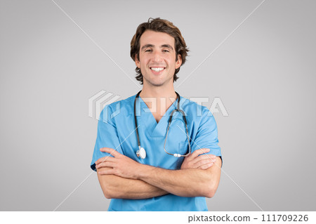Positive confident man nurse, doctor in blue uniform with crossed arms on chest smiling at camera on grey background 111709226