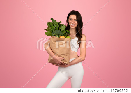 A smiling woman in a white sports outfit confidently holds a grocery bag full of green vegetables and fruits A smiling woman in a white sports outfit confidently holds a grocery bag full of green vegetables and fruits 111709232