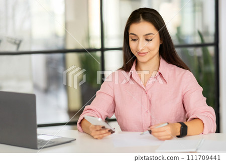 Cheerful millennial arab woman manager typing on phone, at desk with computer Cheerful millennial arab woman manager typing on phone, at desk with computer 111709441