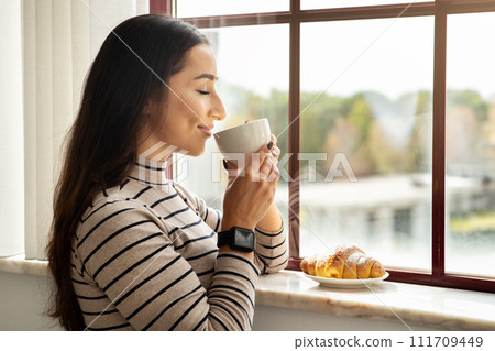 Cheerful young middle eastern lady drink cup of hot coffee, tea with croissant Cheerful young middle eastern lady drink cup of hot coffee, tea with croissant 111709449