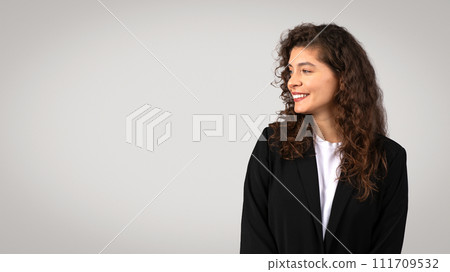 Optimistic businesswoman in black suit looking aside at free space with pleasant smile, grey background, panorama 111709532