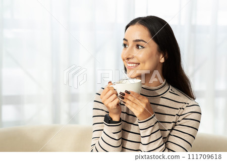 Cheerful young arab woman drink cup of hot coffee, tea in light living room, kitchen interior 111709618