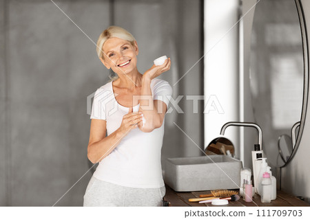 Beautiful Senior Woman Applying Moisturising Cream On Elbow While Standing In Bathroom Beautiful Senior Woman Applying Moisturising Cream On Elbow While Standing In Bathroom 111709703