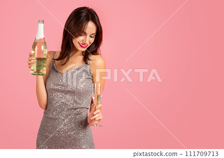 Joyful woman in a glittering silver dress holding a bottle of champagne and a glass 111709713
