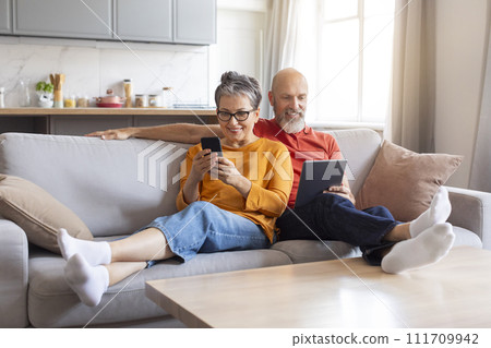 Happy Senior Spouses Using Modern Gadgets While Relaxing On Couch At Home 111709942