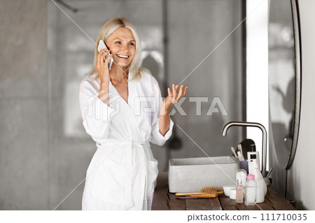 Happy Senior Woman Wearing Bathrobe Talking On Mobile Phone In Modern Bathroom Happy Senior Woman Wearing Bathrobe Talking On Mobile Phone In Modern Bathroom 111710035