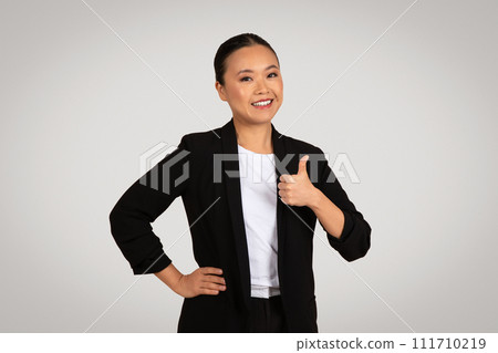 Radiant Asian businesswoman in a chic black suit gives a thumbs up while smiling confidently 111710219