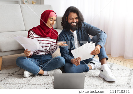 Cheerful young muslim spouses holding financial papers and using laptop at home Cheerful young muslim spouses holding financial papers and using laptop at home 111710251