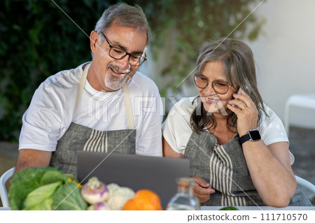 Positive senior caucasian man and woman in aprons use laptop, make phone calls at table 111710570
