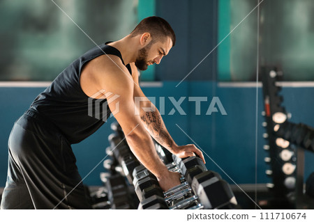 Sport Equipment Concept. Young Male Athlete Taking Dumbbell From Rack At Gym Sport Equipment Concept. Young Male Athlete Taking Dumbbell From Rack At Gym 111710674