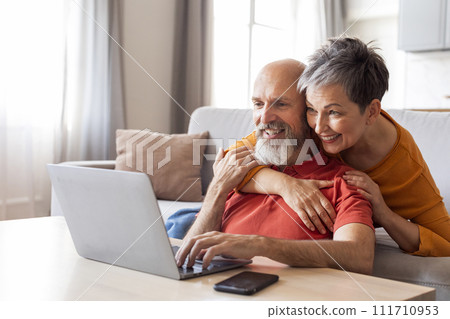 Smiling Senior Spouses Spending Time With Laptop In Living Room Smiling Senior Spouses Spending Time With Laptop In Living Room 111710953