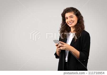 Smiling young businesswoman in formal attire using her smartphone, possibly managing business communications with smile Smiling young businesswoman in formal attire using her smartphone, possibly managing business communications with smile 111710954