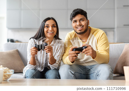 Cheerful Indian Couple At Home Playing Video Games With Joysticks 111710996