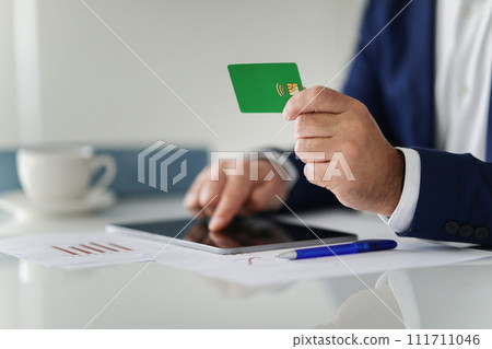 A businessman in a navy suit is making an online payment using a green credit card and a digital tablet A businessman in a navy suit is making an online payment using a green credit card and a digital tablet 111711046