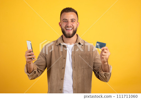 Glad millennial european man with beard show credit card, use phone, celebrate success victory 111711060