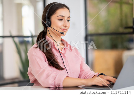Smiling millennial arab woman manager in headset typing on laptop, enjoy work 111711114
