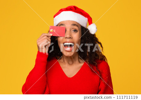 Excited black lady wearing Santa hat, hides eye with credit card, yellow background 111711119