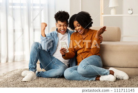Glad excited young black couple winners, with smartphone celebrate win, success, rise fist, sit on floor, good news Glad excited young black couple winners, with smartphone celebrate win, success, rise fist, sit on floor, good news 111711145