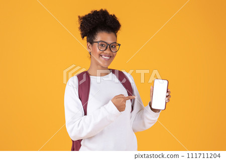 Happy young black woman wearing glasses and backpack pointing at blank smartphone 111711204