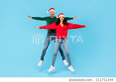Joyful young couple in Santa hats jumping spreading arms, studio 111711244