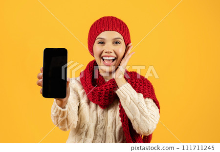 Joyful woman in winter knitwear showcases phone blank screen, studio 111711245