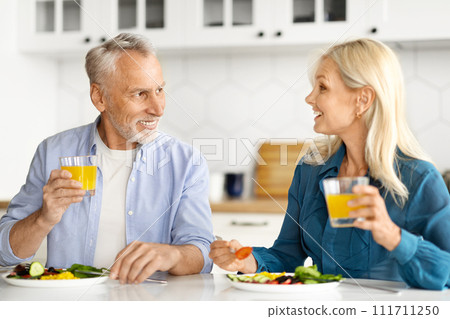Happy Senior Spouses Chatting And Drinking Orange Juice During Breakfast In Kitchen Happy Senior Spouses Chatting And Drinking Orange Juice During Breakfast In Kitchen 111711250
