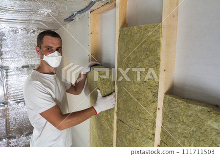 Young worker in respirator insulating rock wool insulation staff in wooden frame for future house walls for cold barrier. Comfortable warm home, economy, construction and renovation concept. Young worker in respirator insulating rock wool insulation staff in wooden frame for future house walls for cold barrier. Comfortable warm home, economy, construction and renovation concept. 111711503