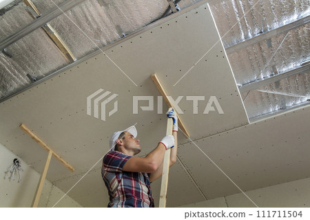 Young worker in protection work gloves fixing wooden holders for drywall suspended ceiling to metal frame insulated with shiny aluminum foil. DIY, do it yourself concept. 111711504