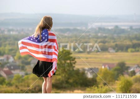 Young pretty american woman with long hair holding waving on wind USA flag on her sholders standing outdoors enjoying warm summer day. 111711550