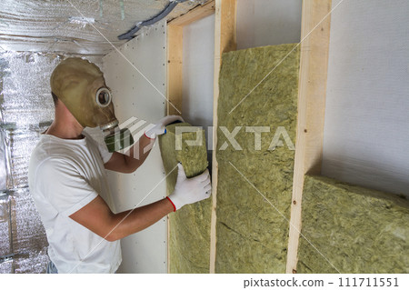 Young muscular worker in gas mask insulating rock wool staff in wooden frame for future house walls for cold and heat barrier. Comfortable warm home, economy, construction and renovation concept. 111711551