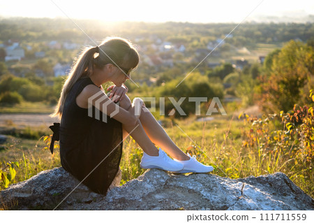 Young depressed woman in black short summer dress sitting on a mountain hill thinking outdoors at sunset. Lonely female contemplating in warm evening in nature. Young depressed woman in black short summer dress sitting on a mountain hill thinking outdoors at sunset. Lonely female contemplating in warm evening in nature. 111711559