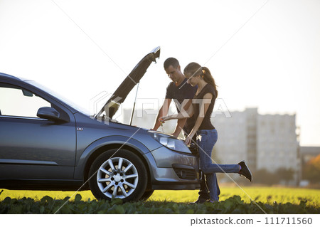 Young couple, handsome man and attractive woman at car with popped hood checking oil level in engine using dipstick on clear sky background. Transportation, vehicles problems and breakdowns concept. Young couple, handsome man and attractive woman at car with popped hood checking oil level in engine using dipstick on clear sky background. Transportation, vehicles problems and breakdowns concept. 111711560