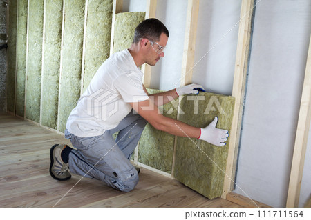 Worker in protective goggles and respirator insulating rock wool insulation in wooden frame for future house walls for cold barrier. Comfortable warm home, economy, construction and renovation concept 111711564