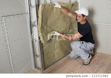 Worker in protective gloves insulating rock wool insulation in wooden frame for future house walls for cold barrier. Comfortable warm home, economy, construction and renovation concept Worker in protective gloves insulating rock wool insulation in wooden frame for future house walls for cold barrier. Comfortable warm home, economy, construction and renovation concept 111711565