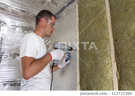 Worker in goggles with screwdriver working on insulation. Drywall on wall beams, insulating rock wool staff in wooden frame. Comfortable warm home, economy, construction and renovation concept. 111711566