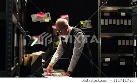 Police officer examining information on files classified records, reading witness statements to build case against culprit. Detective evaluating fingerprints and evidence authenticity. Tripod shot. Police officer examining information on files classified records, reading witness statements to build case against culprit. Detective evaluating fingerprints and evidence authenticity. Tripod shot. 111711755