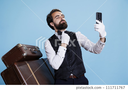 In studio, bellman in uniform takes pictures while using mobile device for selfies and acting foolish against blue background. Classy hotel porter does photos with his phone while being on duty. 111711767