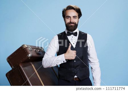 Bellboy gives the thumbs up whilst in studio, looking assured and enthusiastic on camera. Young hotel staff showing gesture of acceptance or consent while donning appropriate attire. Bellboy gives the thumbs up whilst in studio, looking assured and enthusiastic on camera. Young hotel staff showing gesture of acceptance or consent while donning appropriate attire. 111711774