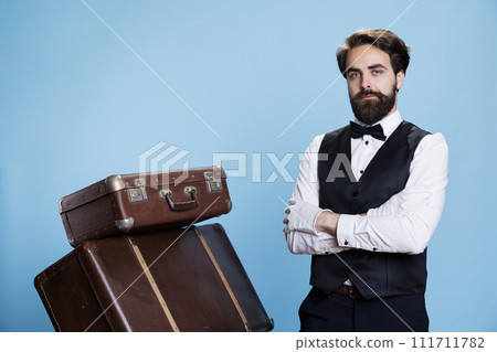 Formal hotel worker posing with luggage, wearing formal suit and white gloves over blue background. Male bellhop standing next to pile of tolley bags and suitcases, hospitality industry. Formal hotel worker posing with luggage, wearing formal suit and white gloves over blue background. Male bellhop standing next to pile of tolley bags and suitcases, hospitality industry. 111711782