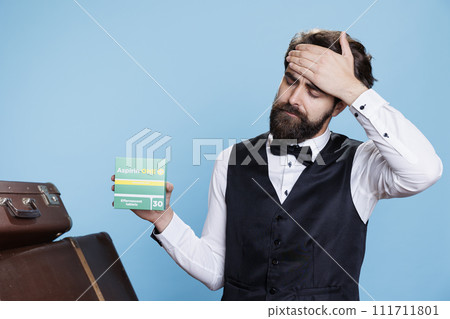 Doorman displays box of pain relievers for headache while posing against blue background. Male bellhop holding medication pack and seeking to ease migraine with medical products in studio. 111711801