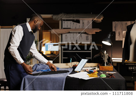 Expert suitmaker stiching together bespoken business suit, looking online for inspiration. Skilled couturier working in professional atelier shop, creating premium quality custom made clothing piece 111711808