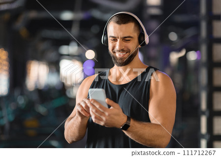 Handsome Muscular Male Athlete In Wireless Headphones Using Mobile Phone At Gym Handsome Muscular Male Athlete In Wireless Headphones Using Mobile Phone At Gym 111712267