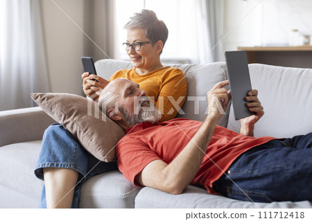 Happy Senior Spouses Relaxing With Modern Gadgets On Couch At Home Happy Senior Spouses Relaxing With Modern Gadgets On Couch At Home 111712418