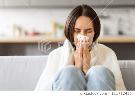 Sick Young Woman Blowing Nose In Paper Tissue While Sitting On Couch 111712435