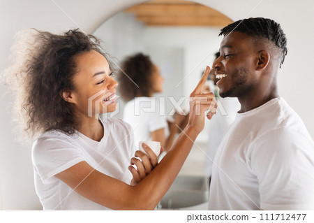 Playful Black Millennial Lady Applying Moisturizer On Husband's Nose Indoor 111712477
