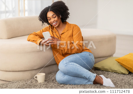Smiling African American woman in a mustard shirt and blue jeans sitting comfortably on the floor 111712478