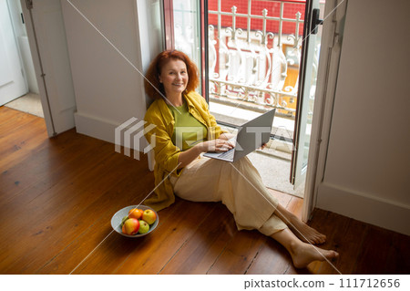 Happy senior lady with laptop computer sitting on floor by opened window 111712656