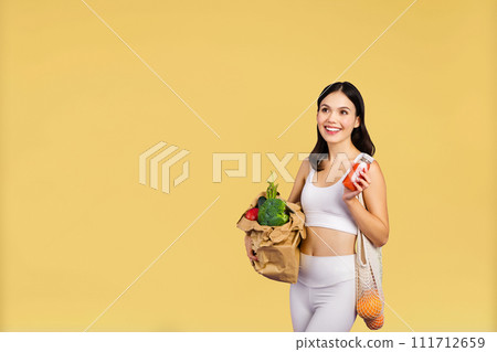 Smiling young woman in sportswear holding paper bag with vegetables and smoothie bottle on yellow studio background 111712659